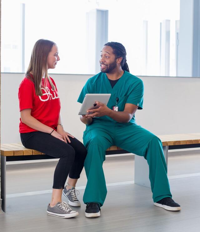 A healthcare professional sits on a bench, holding a tablet and showing it to an Ohio State student